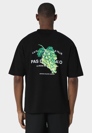Mand iført sort T-shirt med stor grøn drueklase grafik og hvid fransk tekst på bagsiden, stående mod en ensfarvet baggrund.