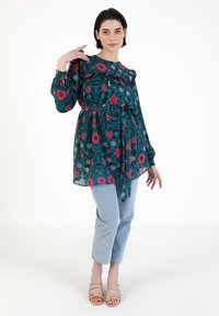 Top w kwiatowy wzór w kolorze teal z falbaniastym dekoltem, wiązanym w pasie i długimi rękawami, w połączeniu z jasnoniebieskimi dżinsami i beżowymi sandałami na ramiączkach.