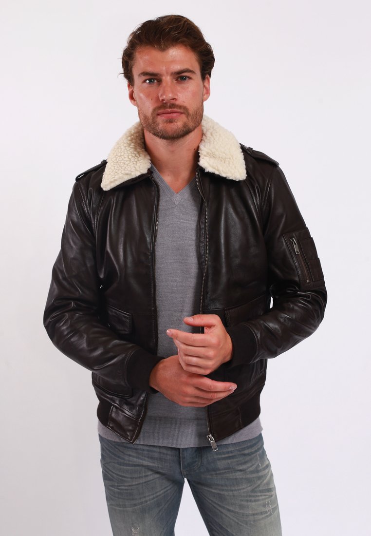 Brown leather bomber jacket with a shearling collar. Features front pockets, zip closure, and ribbed cuffs. Worn over a gray v-neck sweater.
