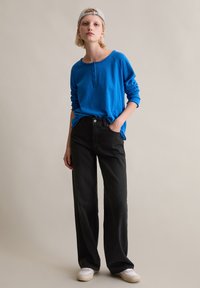 Niebieska koszula henley z długim rękawem, czarne spodnie o szerokich nogawkach, białe trampki. Model stoi z jedną ręką w kieszeni na neutralnym tle.