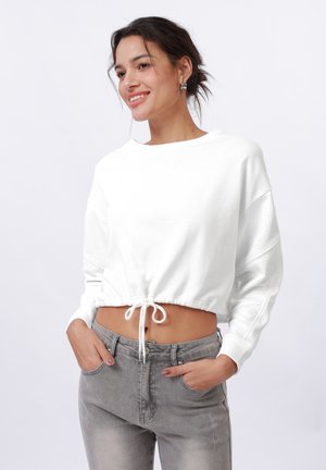 Felpa bianca corta con maniche ampie e orlo con coulisse. Abbinata a jeans grigi, che mostrano una vestibilità rilassata e una superficie liscia.