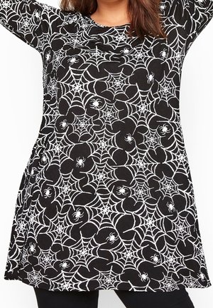 Donna che indossa un vestito nero a maniche lunghe con motivi bianchi di ragnatela e ragno, in piedi con le braccia alzate sopra la testa.