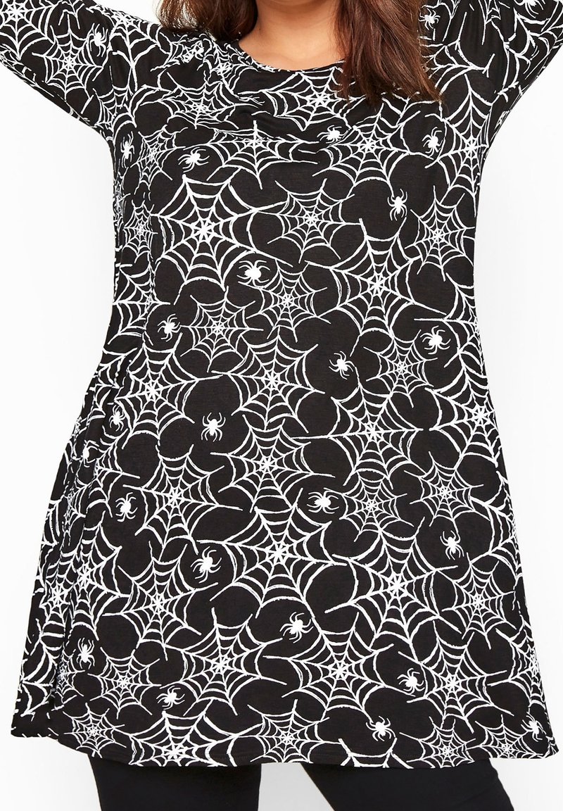 Femme portant une robe noire à manches longues avec des motifs de toile d'araignée blanche et d'araignée, debout les bras levés au-dessus de la tête.