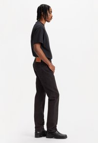 Hombre con cabello trenzado de perfil, viste camiseta negra, jeans oscuros con un parche marrón y zapatos de cuero negros, frente a un fondo blanco.