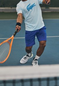 Light blue t-shirt with a blue crocodile logo, paired with textured purple shorts. White and black tennis shoes with green accents. Tennis racket held.