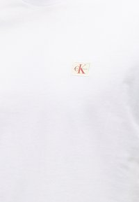 Λευκό ύφασμα με ένα μικρό μπεζ τετράγωνο ετικέτα που έχει κόκκινο "cK" και αχνό κείμενο "Calvin Klein" στην περιοχή του άνω στήθους.