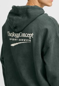 Felpa di colore verde scuro con una texture morbida, caratterizzata da testo bianco "TheJoggConcept" e "SPORT SERVICE" stampato sul retro. Include un cappuccio.