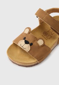 Καφέ δερμάτινο σανδάλι με σχεδιασμό προσώπου αρκούδας, αυτιά και ραμμένες λεπτομέρειες. Ρυθμιζόμενο λουράκι και υφασμένο πέλμα για άνεση.