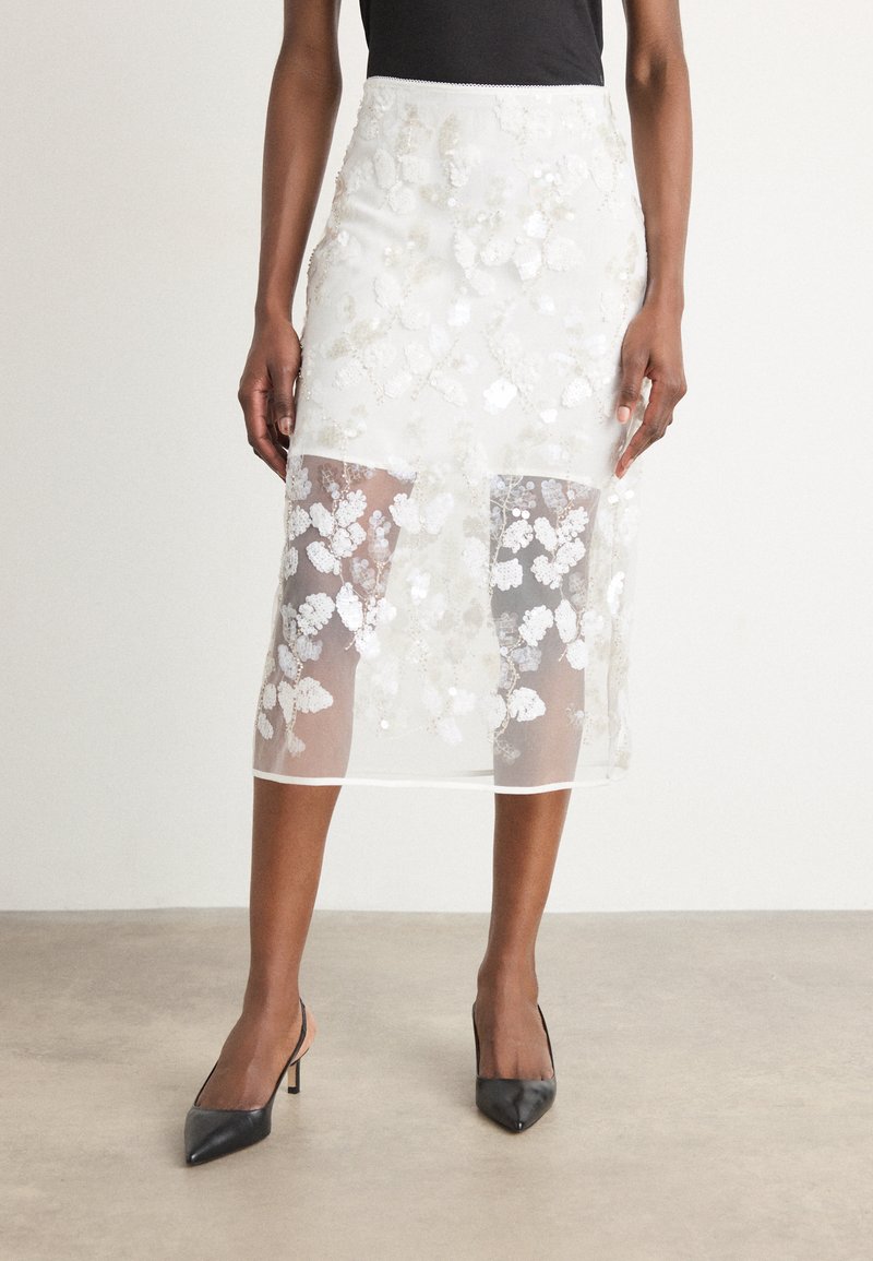 Falda midi blanca con bordado floral y lentejuelas. Superposición transparente y detalles en el dobladillo. Combinada con zapatos de tacón negro de punta afilada.
