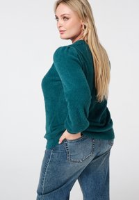 Turfkleurige, pluizige trui met geplooide lange mouwen, gecombineerd met lichtblauwe spijkerbroek. De trui heeft een ontspannen pasvorm en een gestructureerde stof.