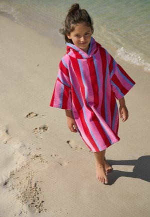Barn går barfodet på sandet strand iført en rød, pink og blåstribet hættetrøje-poncho nær vandkanten.