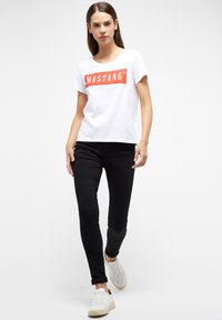 Camiseta blanca de manga corta con un prominente logo rectangular rojo "MUSTANG", combinada con pantalones negros ajustados y zapatillas blancas.