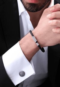 Bracciale in pelle intrecciata nera con accenti in metallo argentato indossato su un polso, abbinato a una camicia bianca e a una giacca da suit nera.