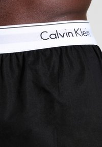 Crne bokserske gaće Calvin Klein s bijelim elastičnim pojasom na kojem je naziv brenda ispisan crnim slovima i strukturiranom tkaninom.