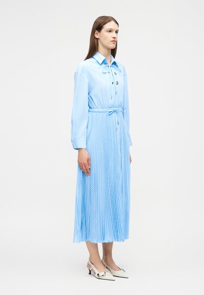 Γυναίκα που στέκεται φορώντας ένα ανοιχτό μπλε midi φόρεμα με μακριά μανίκια, πιέτες και λευκές ρίγες, με δέσιμο στη μέση, καθώς και λευκά παπούτσια με χαμηλό τακούνι και φιόγκους.