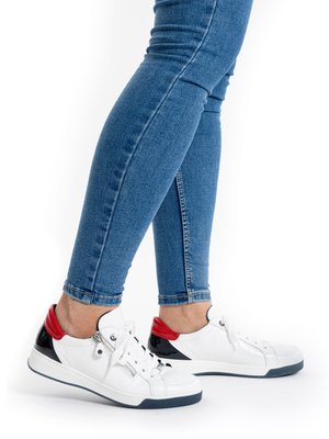 Legs in blue skinny jeans wearing white sneakers with red and black accents on a white background.