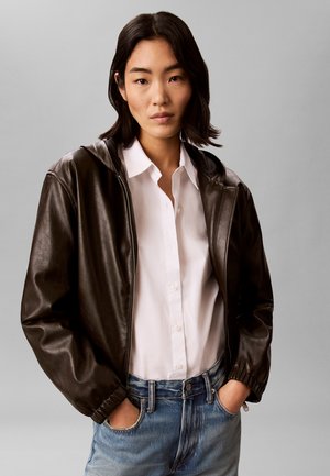 Jeune femme portant une veste en cuir marron foncé sur une chemise blanche à boutons et un jean bleu, mains dans les poches, expression neutre.
