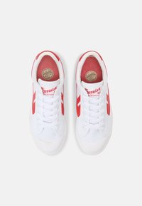 Baskets en toile blanches avec des accents rouges, dotées d'une pointe ronde, de lacets fins et d'un logo sur la languette. La semelle est lisse et légèrement texturée.