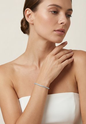 Donna con capelli raccolti che indossa un top bianco senza spalline, che tocca delicatamente la spalla, mostrando un bracciale tennis d'argento al polso.