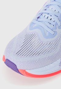 Zapatilla de correr de color púrpura claro con parte superior de malla texturizada, que presenta una suela rosa y púrpura y cordones azules. Elementos de diseño destacables que permiten una buena transpiración.
