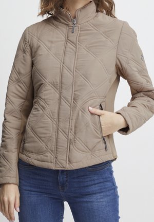 Femme portant une veste matelassée beige avec fermeture éclair frontale et poches zippées, associée à un jean bleu, main dans la poche, sur fond uni.