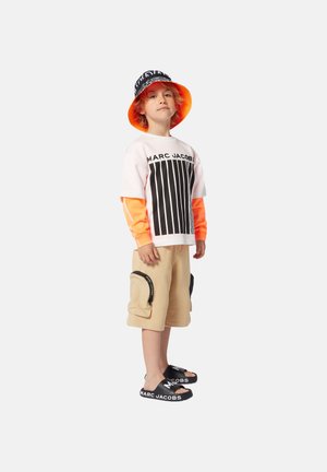 T-shirt bianco con strisce nere e testo "MARC JACOBS", maniche arancioni, pantaloni cargo beige, ciabatte nere con logo e cappello a secchiello coordinato.