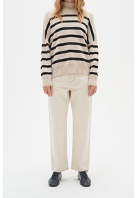 Person trägt beigen weit geschnittenen Hosen, einen beigen Pullover mit schwarzen horizontalen Streifen und schwarze Schnürschuhe, steht vor einem weißen Hintergrund.