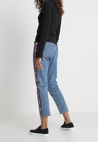 Jeans di denim azzurro chiaro con strisce laterali rosse e nere. I jeans sono di taglio largo e cropped, abbinati a sneakers nere.