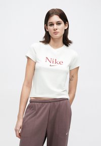 Νέος με καστανά μαλλιά που φτάνουν μέχρι τους ώμους, ντυμένος με λευκή κοντή μπλούζα Nike και χαλαρές καφέ φόρμες, στέκεται μπροστά σε απλή πλάτη.