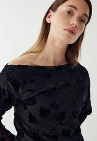 Fekete, vállnál nyitott blúz textúrált virágmintával, áttetsző anyagból és sima felülettel. Diszkrét fénylés jellemzi, laza fazonnal és hosszú ujjakkal.