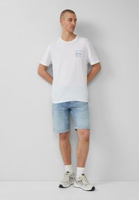 T-shirt en coton blanc avec imprimé graphique bleu, associé à un short en denim clair. Le modèle porte des chaussettes blanches et des baskets grises. Design simple, coupe décontractée.
