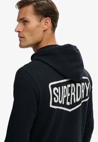 Sudadera con capucha negra hecha de tela suave, con un gran logo blanco "SUPERDRY" en la espalda. Incluye una capucha espaciosa y puños acanalados.