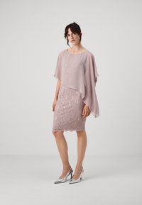 Krátky, splývavý blušový chiffon top nad priliehavými čipkovými šatami s akcentmi z flitrov; obsahuje scalloped lem. Modelka má na sebe strieborné vysoké topánky.