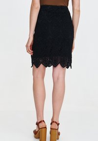 Schwarzer Spitzenrock mit gewelltem Saum, der aufwendige Blumenmuster und eine figurbetonte Passform aufweist. Trägt dazu braune Blockabsatzsandalen.