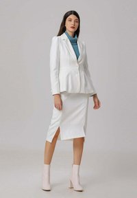 Femme se tient debout portant un blazer blanc et une jupe assortie avec des fentes sur le devant, un pull à col roulé bleu et des bottines blanches sur un fond uni.