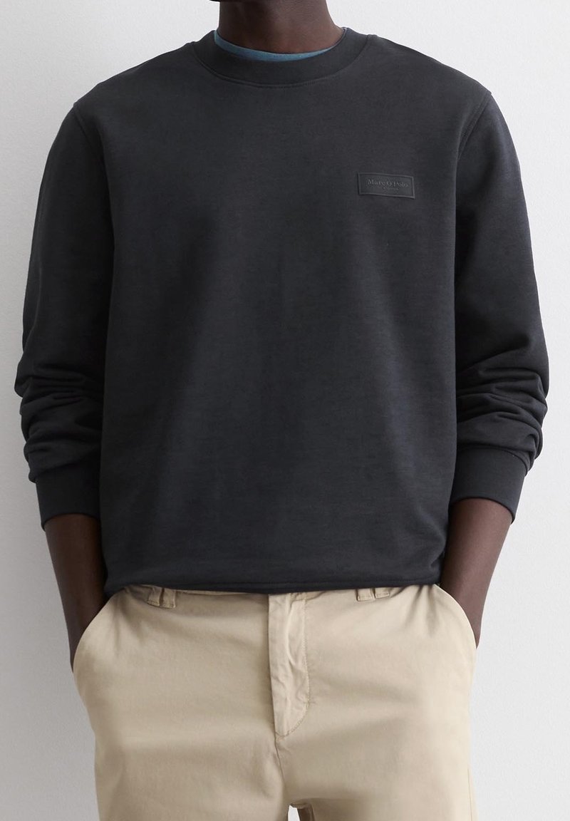 Sweat-shirt noir en tissu doux avec un col rond et des manches à bords-côtes, doté d'un petit patch logo rectangulaire sur la poitrine.
