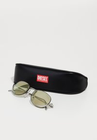Ochelari de soare cu lentile verzi, rame metalice argintii și formă ovală. Însoțiți de un etui din piele neagră cu un logo roșu Diesel.