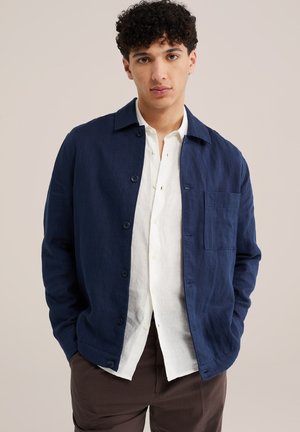 Giovane uomo con capelli ricci che indossa una giacca blu navy con bottoni, camicia bianca e pantaloni marroni a righe sottili, in piedi davanti a uno sfondo semplice.