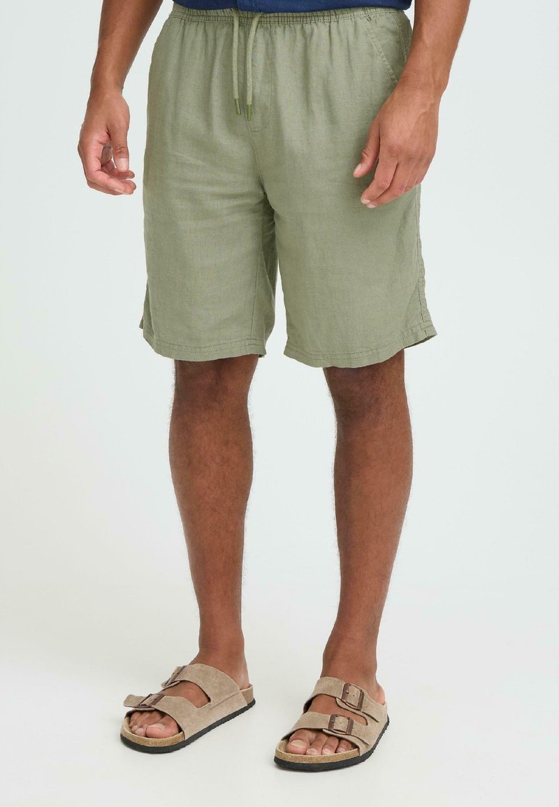 Shorts en lin vert olive avec une taille élastique et un cordon de serrage, dotés de poches latérales et d'une coupe décontractée. Associés à des sandales beiges.
