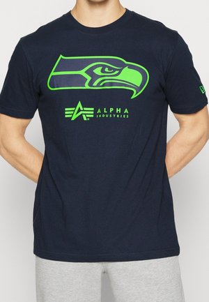 Námořnická modrá bavlněná tričko s velkým zeleným logem Seattle Seahawks a textem "ALPHA INDUSTRIES" v jasně zelené barvě. Krátké rukávy, kulatý výstřih.