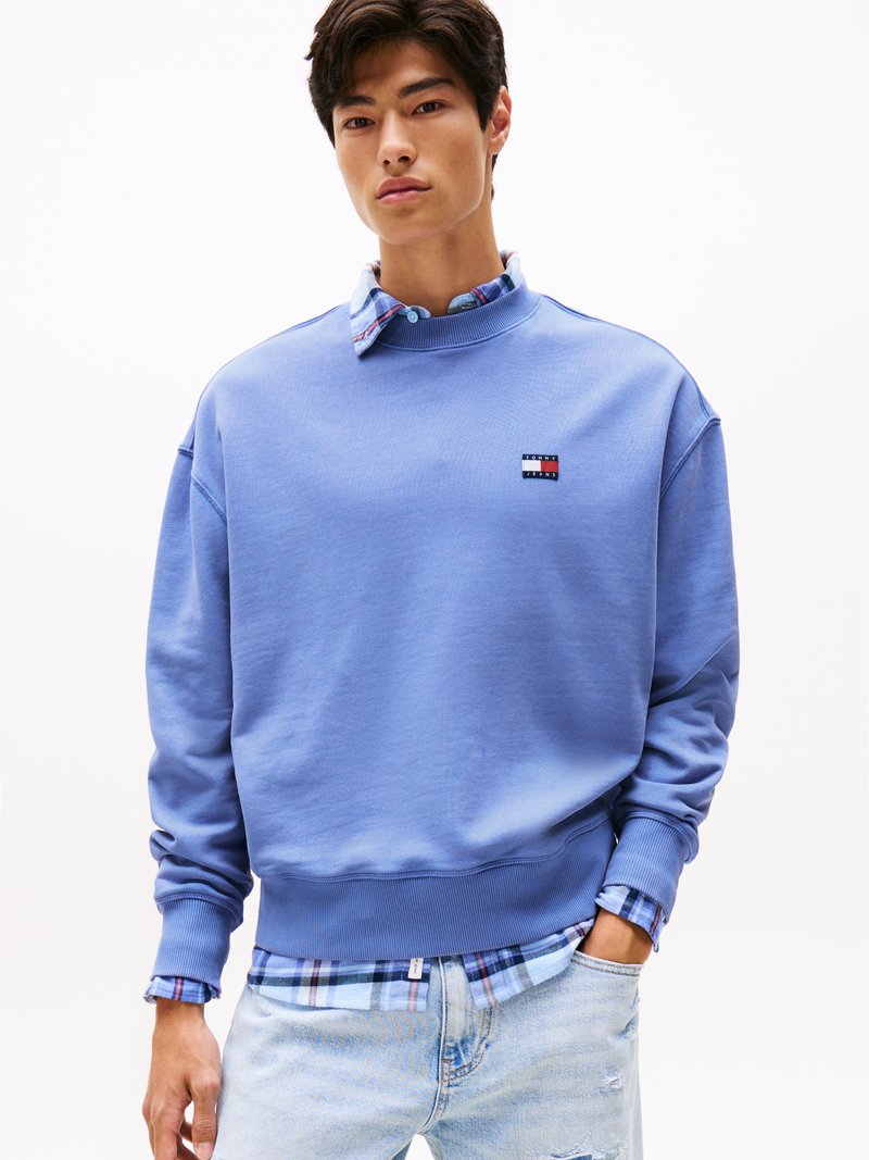 Sweatshirt azul em algodão com punhos canelados, apresentando um pequeno logotipo. Por baixo, uma camisa xadrez em tons de azul exibe um design em camadas.