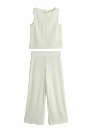 Femeie cu păr scurt maro poartă un top fără mâneci tricotat crem și o fustă, ajustând materialul în talie, pe un fundal alb simplu.