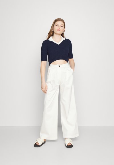 Sweater cropped azul-marinho com mangas curtas e colarinho branco, combinado com calças largas brancas com detalhe de botão e sandálias pretas.