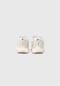 Sneakers Nike z jasnobeżową i szaro-siatkowaną cholewką, teksturowanymi akcentami, wyraźnym wsparciem pięty oraz czarnym logotypem na pięcie.