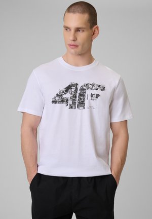 Mężczyzna o krótkich włosach, ubrany w biały t-shirt z grafiką i czarne spodnie, ręce w kieszeniach, patrzący na bok na szarym tle.