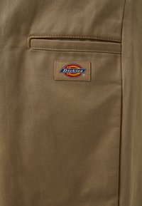 Smeđi radni hlače od tkanine s remenom, imaju zakrpe džep i šarenu oznaku Dickies ušivenu na materijal. Glatka tekstura, izdržljiv dizajn.