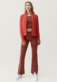 Punainen vuorattu takki pystykauluksella, yhdistettynä ruskeaan istuvaan puseroon ja yhteensopiviin leveälahkeisiin housuihin. Valkoiset lenkkarit korostavat asua.