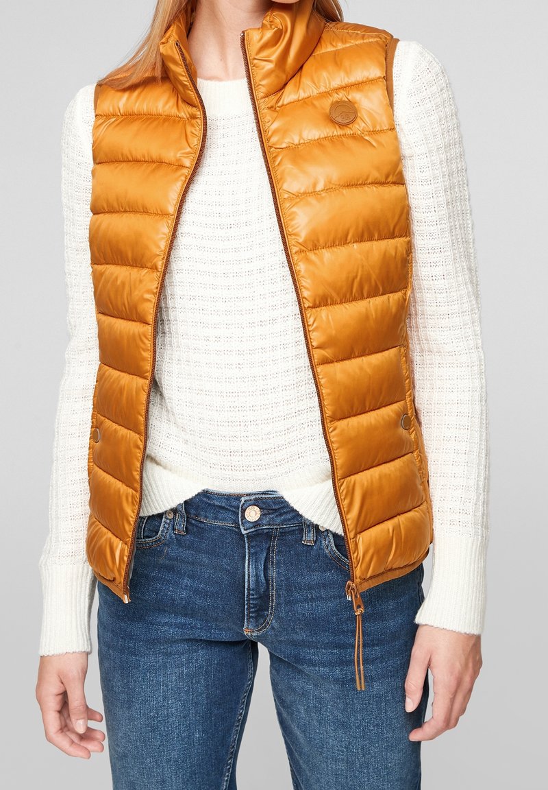 Personne portant une doudoune jaune moutarde sur un pull en tricot blanc avec un jean bleu sur un fond clair uni.