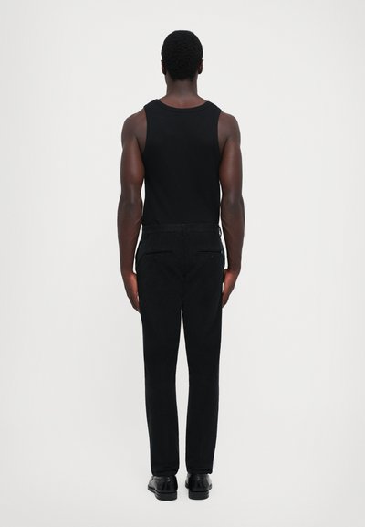 Hombre de pie de espaldas, vistiendo una camiseta sin mangas negra, pantalones negros y zapatos negros, contra un fondo claro y liso.