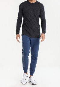 Svart långärmad texturerad skjorta i kombination med blå joggers som har en stor grå "Under Armour"-logotyp längs vänster ben. Vita sneakers.